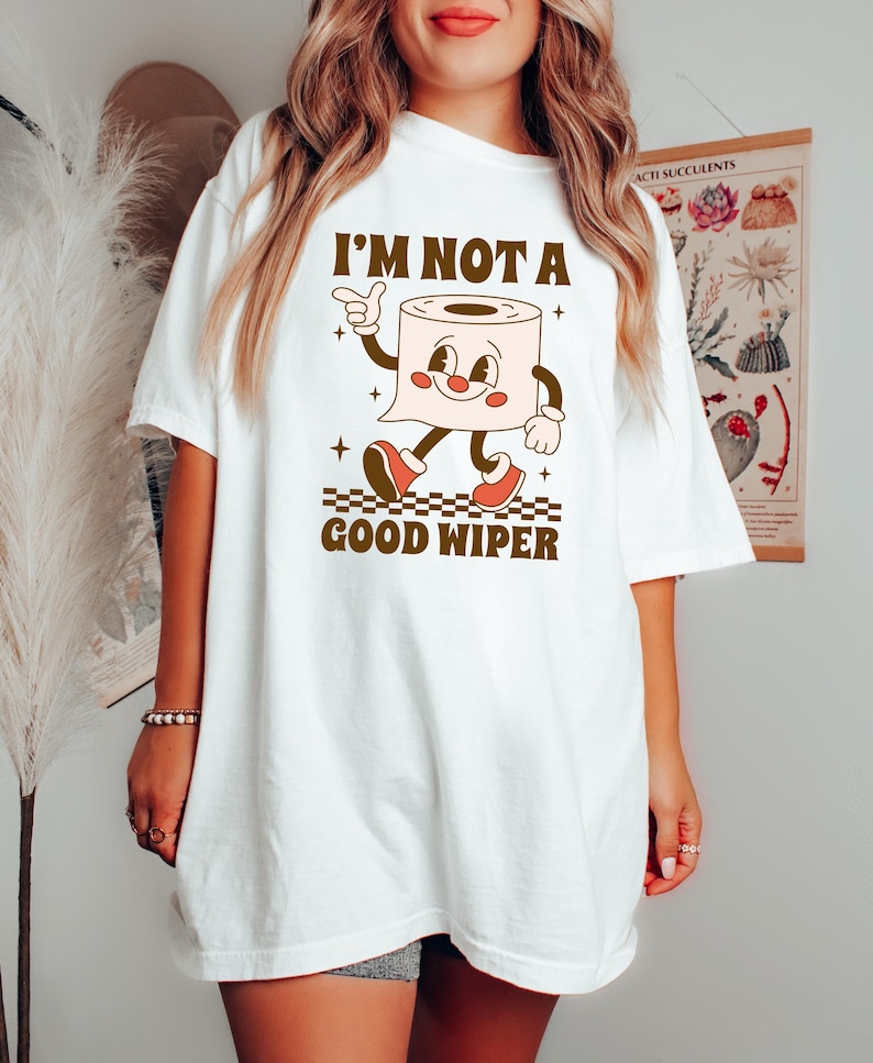 May include: White t-shirt with a retro cartoon toilet paper roll character. The character has a smiling face, arms, and legs, wearing red shoes. The text "I'M NOT A GOOD WIPER" is printed above and below the character in brown.