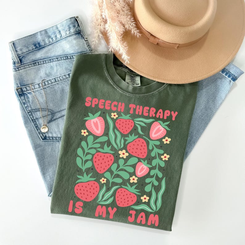 May include: An olive green t-shirt with the text "SPEECH THERAPY IS MY JAM" in pink. The shirt features a strawberry design with pink strawberries and green leaves. A pair of light blue jeans and a tan hat are also in the image.