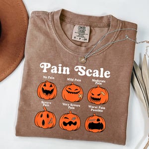 May include: A brown t-shirt with the text "Pain Scale" and illustrations of jack-o'-lanterns representing different levels of pain. The shirt has a crew neck and a small tag at the top. A silver necklace with a pendant is also visible.