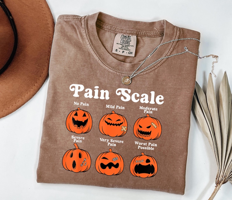May include: A brown t-shirt with the text "Pain Scale" and illustrations of jack-o'-lanterns representing different levels of pain. The shirt has a crew neck and a small tag at the top. A silver necklace with a pendant is also visible.