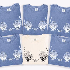 May include: Six t-shirts are shown, five in blue and one in cream. Each shirt has a white graphic of two seashells with the text "Bachelorette" and "March 2026". The shirts are folded and arranged on a white surface.