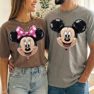 Peut inclure: Un t-shirt marron avec un motif de ballon Minnie Mouse et un t-shirt gris avec un motif de ballon Mickey Mouse. Les deux v&ecirc;tements pr&eacute;sentent les visages embl&eacute;matiques des personnages sous forme de ballons ronds et brillants avec un fil rouge.