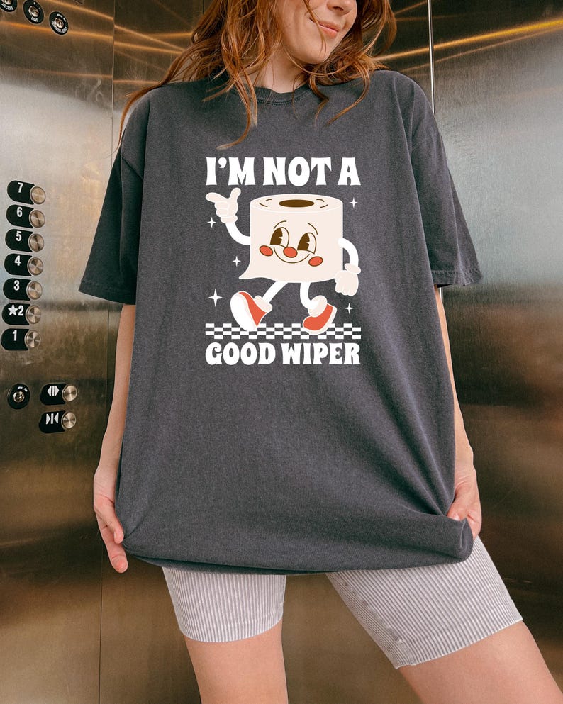 May include: Dark gray t-shirt with the text "I'M NOT A GOOD WIPER" and a cartoon toilet paper roll character. The character has a smiling face, arms, and red shoes. The shirt is a relaxed fit.