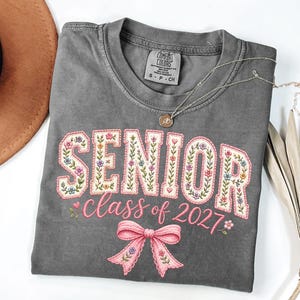 Può includere: Una t-shirt grigio antracite a maniche corte con la parola SENIOR in grandi lettere stampatello riempite di motivi floreali, il testo class of 2027 in corsivo rosa e un fiocco rosa. La maglietta ha un'etichetta sul collo Comfort Colors.