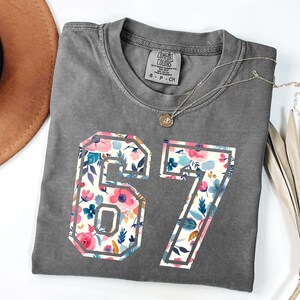 May include: A gray Comfort Colors t-shirt with the number "67" in a floral design. The flowers are pink, blue, and white. A gold necklace with a pendant is on the shirt.