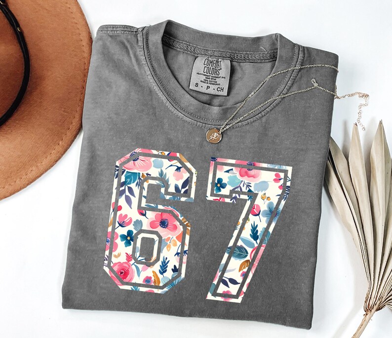 May include: A gray Comfort Colors t-shirt with the number "67" in a floral design. The flowers are pink, blue, and white. A gold necklace with a pendant is on the shirt.