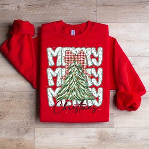 May include: A red sweatshirt featuring a Christmas tree design. The words "Merry Christmas" are displayed in a festive font, with a plaid bow at the top of the tree. The sleeves are long and the cuffs are ribbed.