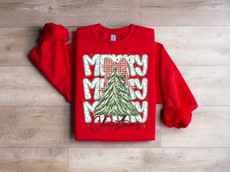 May include: A red sweatshirt featuring a Christmas tree design. The words "Merry Christmas" are displayed in a festive font, with a plaid bow at the top of the tree. The sleeves are long and the cuffs are ribbed.