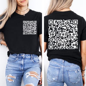 May include: A black short-sleeved t-shirt featuring a white QR code graphic on the front left chest and a larger white QR code graphic printed on the back. The shirt is paired with light blue distressed denim jeans with visible rips.