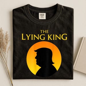 May include: A black t-shirt featuring the yellow text THE LYING KING above a circular graphic. The graphic contains a black silhouette of a person's head in profile against a gradient orange and yellow sun-like background. The shirt has a neck label.
