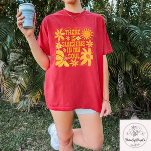 May include: Red t-shirt with the text "THERE IS SUNSHINE IN MY SOUL" in yellow floral design. The shirt is a short-sleeved, casual style. The person is holding a light blue collapsible cup.