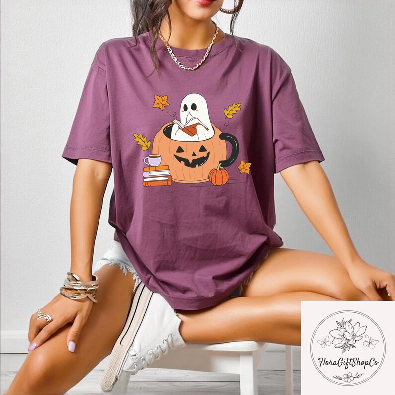 May include: A purple t-shirt featuring a Halloween-themed graphic. The design includes a ghost reading inside a jack-o'-lantern mug, with books, a teacup, and a pumpkin. The shirt is a short-sleeved, casual style.