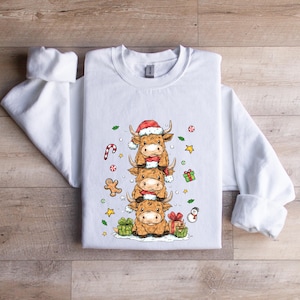 May include: White sweatshirt with a festive design of three cartoon Highland cows, each wearing a Santa hat and scarf. The design includes Christmas decorations like candy canes, gingerbread men, and wrapped gifts. The sweatshirt is on a wooden surface.