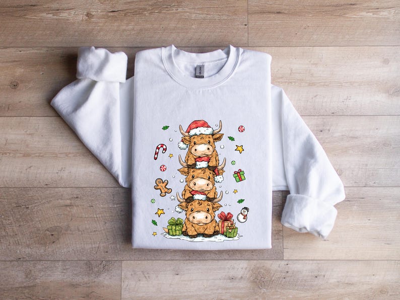 May include: White sweatshirt with a festive design of three cartoon Highland cows, each wearing a Santa hat and scarf. The design includes Christmas decorations like candy canes, gingerbread men, and wrapped gifts. The sweatshirt is on a wooden surface.