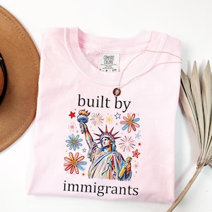 May include: A light pink t-shirt featuring a graphic of the Statue of Liberty surrounded by colorful flowers and fireworks. The text "built by immigrants" is printed in black above and below the graphic. The shirt has a Comfort Colors label.