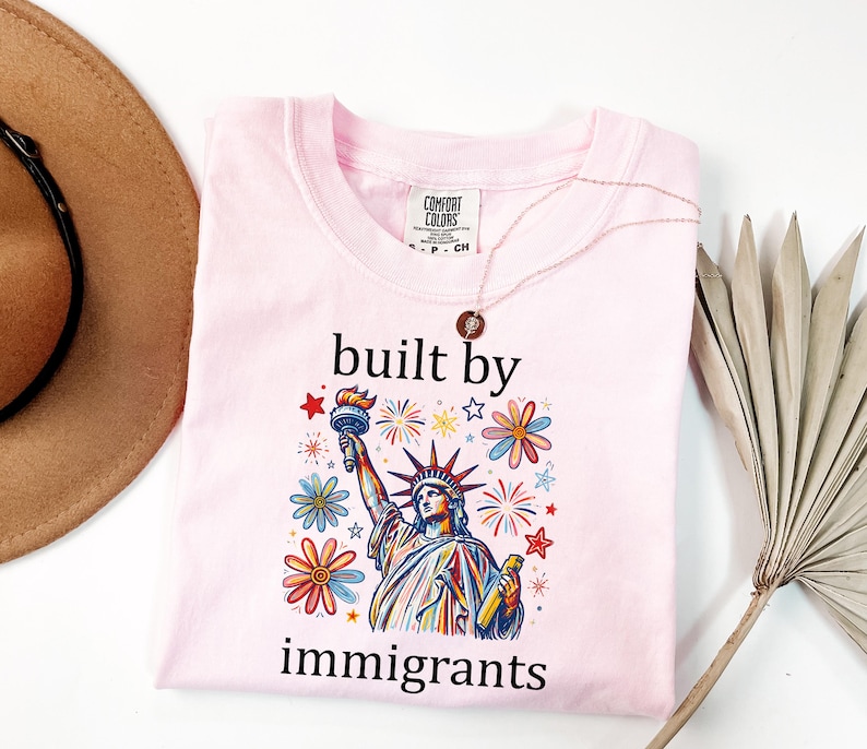 May include: A light pink t-shirt featuring a graphic of the Statue of Liberty surrounded by colorful flowers and fireworks. The text "built by immigrants" is printed in black above and below the graphic. The shirt has a Comfort Colors label.