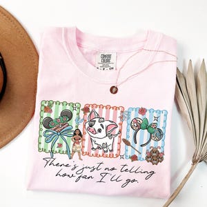 May include: A light pink t-shirt featuring a graphic print of a character, a pig, and two sets of mouse ears within three rectangular frames. Below the graphics, the text reads, "There's just no telling how far I'll go." The shirt has a Comfort Colors label.