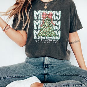 May include: Dark gray t-shirt with a Christmas tree graphic. The design features the words "Merry Merry Merry Christmas" in a stacked layout, with a red and white plaid bow and a decorated Christmas tree.