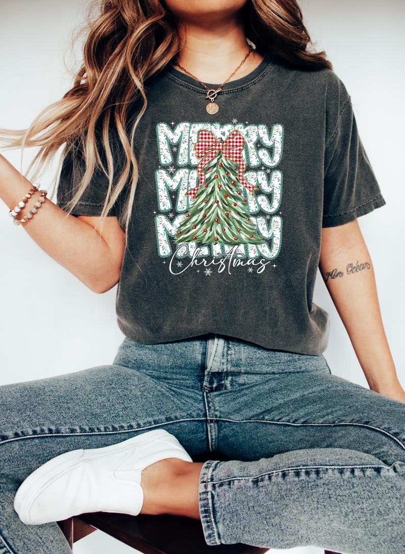 May include: Dark gray t-shirt with a Christmas tree graphic. The design features the words "Merry Merry Merry Christmas" in a stacked layout, with a red and white plaid bow and a decorated Christmas tree.
