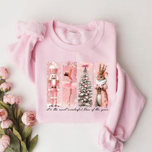 May include: A light pink sweatshirt featuring a festive design with a nutcracker, ballerina, Christmas tree, and a mouse in a coat. The words "It's the most wonderful time of the year" are printed at the bottom.