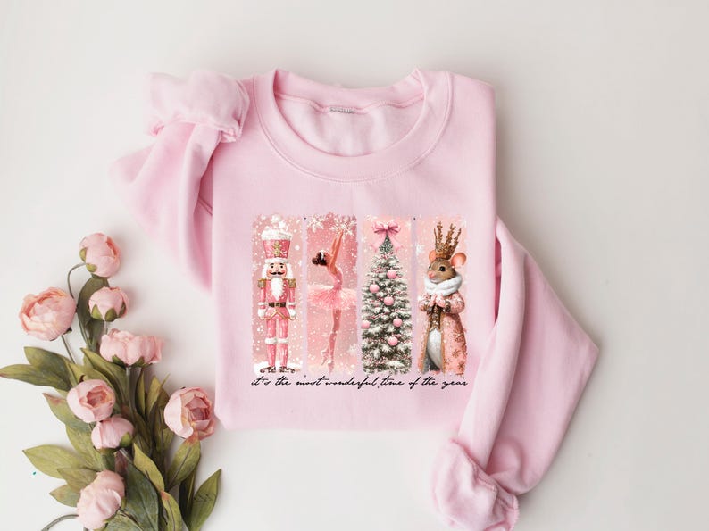 May include: A light pink sweatshirt featuring a festive design with a nutcracker, ballerina, Christmas tree, and a mouse in a coat. The words "It's the most wonderful time of the year" are printed at the bottom.