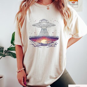 Op de afbeelding: Cr&egrave;mekleurig T-shirt met een gepixeld grafisch ontwerp van een UFO die licht uitstraalt op een zonsondergang. Het ontwerp bevat een gedetailleerde weergave van het ruimteschip en het landschap eronder. Het shirt is een comfortabel, casual kledingstuk.