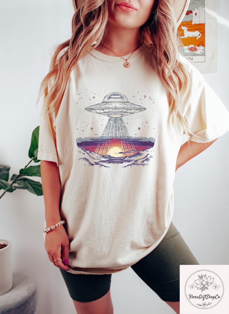 Op de afbeelding: Cr&egrave;mekleurig T-shirt met een gepixeld grafisch ontwerp van een UFO die licht uitstraalt op een zonsondergang. Het ontwerp bevat een gedetailleerde weergave van het ruimteschip en het landschap eronder. Het shirt is een comfortabel, casual kledingstuk.