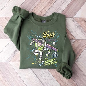 May include: Green crewneck sweatshirt featuring a graphic of Buzz Lightyear from Toy Story. The design includes the text "Buzz Lightyear" and "To Infinity and Beyond" with a space-themed background. The sweatshirt is made of a soft material.