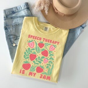 May include: A light yellow t-shirt with the text "SPEECH THERAPY IS MY JAM" in pink. The shirt features a strawberry design with pink strawberries and green leaves. A pair of blue jeans, a tan hat, and a necklace are also in the image.