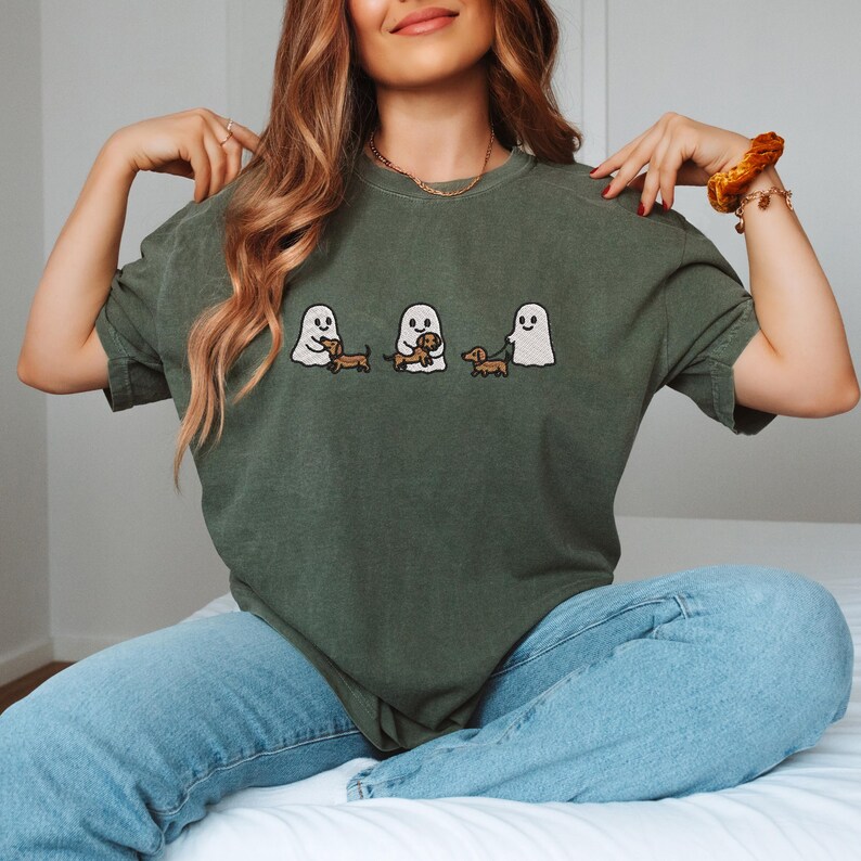 May include: Olive green t-shirt featuring embroidered ghosts with dachshund dogs. The design includes three ghosts, each interacting with a dachshund. The shirt is a relaxed fit and has short sleeves.
