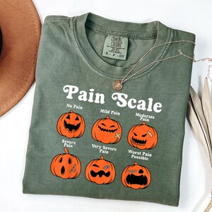 May include: Olive green t-shirt with the text "Pain Scale" and illustrations of jack-o'-lanterns depicting different levels of pain. A gold necklace is draped across the shirt. The shirt has a Comfort Colors tag.