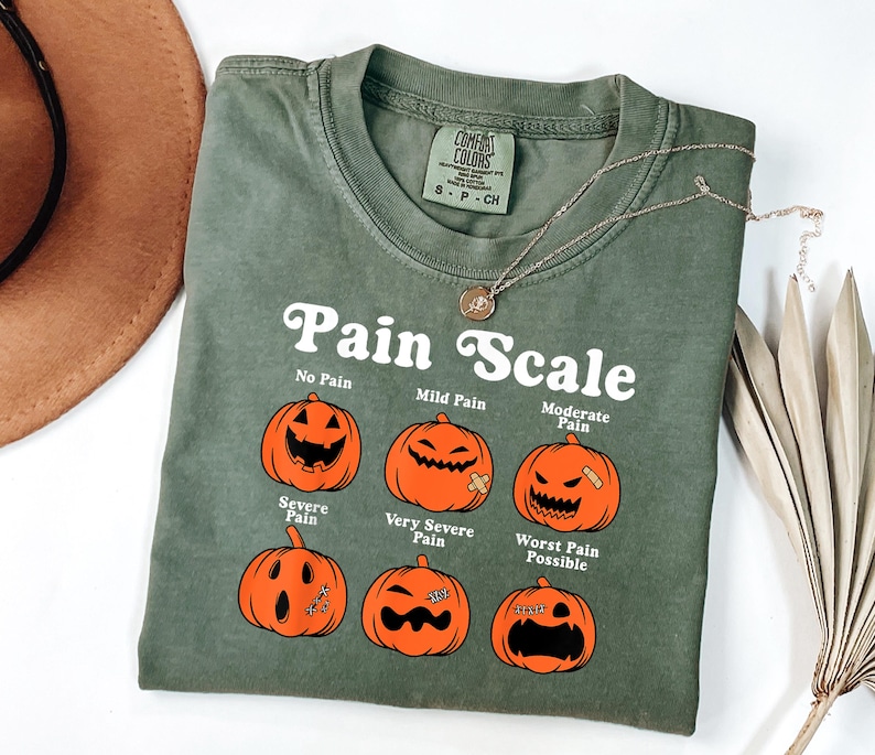 May include: Olive green t-shirt with the text "Pain Scale" and illustrations of jack-o'-lanterns depicting different levels of pain. A gold necklace is draped across the shirt. The shirt has a Comfort Colors tag.