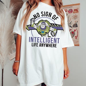 May include: White t-shirt featuring a graphic of Buzz Lightyear with the text "NO SIGN OF INTELLIGENT LIFE ANYWHERE." The Buzz Lightyear graphic is in green, purple, and blue. The text is in black and purple.