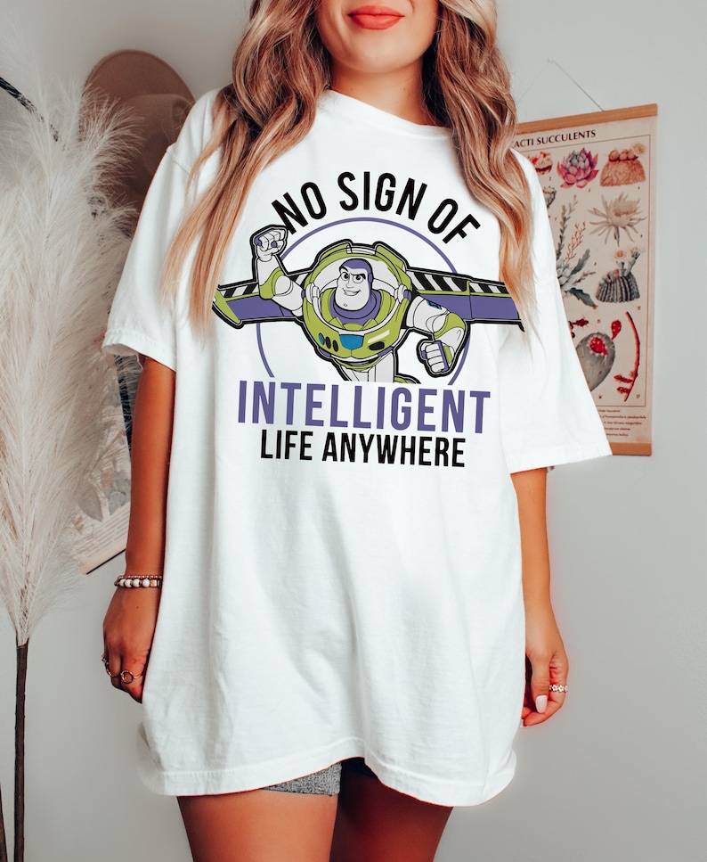 May include: White t-shirt featuring a graphic of Buzz Lightyear with the text "NO SIGN OF INTELLIGENT LIFE ANYWHERE." The Buzz Lightyear graphic is in green, purple, and blue. The text is in black and purple.