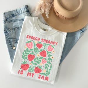 May include: White t-shirt with the text "SPEECH THERAPY IS MY JAM" in pink. The shirt features a strawberry design with green leaves and yellow flowers. The shirt is paired with blue jeans and a tan hat.
