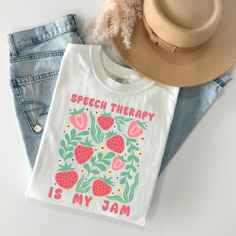 May include: White t-shirt with the text "SPEECH THERAPY IS MY JAM" in pink. The shirt features a strawberry design with green leaves and yellow flowers. The shirt is paired with blue jeans and a tan hat.