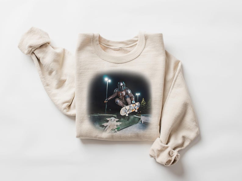 Op de afbeelding: Cr&egrave;mekleurige sweatshirt met een afbeelding van een personage in harnas dat skateboardt met een klein groen wezen. De afbeelding is 's nachts met heldere lichten. De sweatshirt is gemaakt van een zacht materiaal.