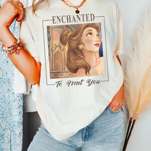 May include: A cream-colored t-shirt featuring a rectangular graphic print of a brown-haired character with a yellow hair ribbon. The text above the graphic reads ENCHANTED and the text below reads To Meet You in a dark brown script font.