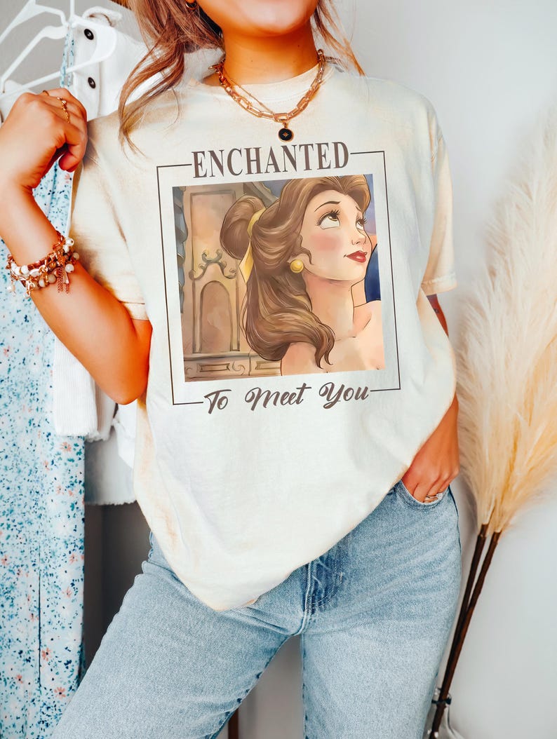May include: A cream-colored t-shirt featuring a rectangular graphic print of a brown-haired character with a yellow hair ribbon. The text above the graphic reads ENCHANTED and the text below reads To Meet You in a dark brown script font.