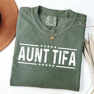 May include: An olive green t-shirt with the text "AUNT TIFA" in white block letters. The shirt has a crew neck and short sleeves. A gold necklace with a pendant is draped across the shirt. A brown hat is in the upper left corner.