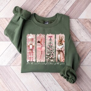 May include: Green sweatshirt with a festive graphic. The graphic features a nutcracker, ballerina, Christmas tree, and mouse in a pink and white color scheme. The text "It's the most wonderful time of the year" is also included.