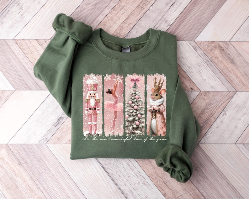 May include: Green sweatshirt with a festive graphic. The graphic features a nutcracker, ballerina, Christmas tree, and mouse in a pink and white color scheme. The text "It's the most wonderful time of the year" is also included.