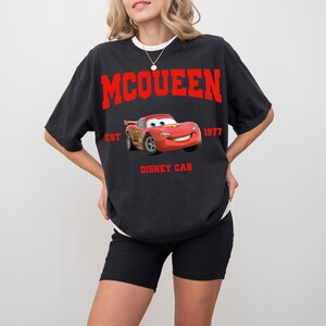 Peut inclure: T-shirt noir avec un motif de Flash McQueen du film Disney Cars. Le motif comprend le nom "McQueen" en lettres rouges, le texte "EST 1977" et "DISNEY CAR". Le t-shirt a un col rond et des manches courtes.