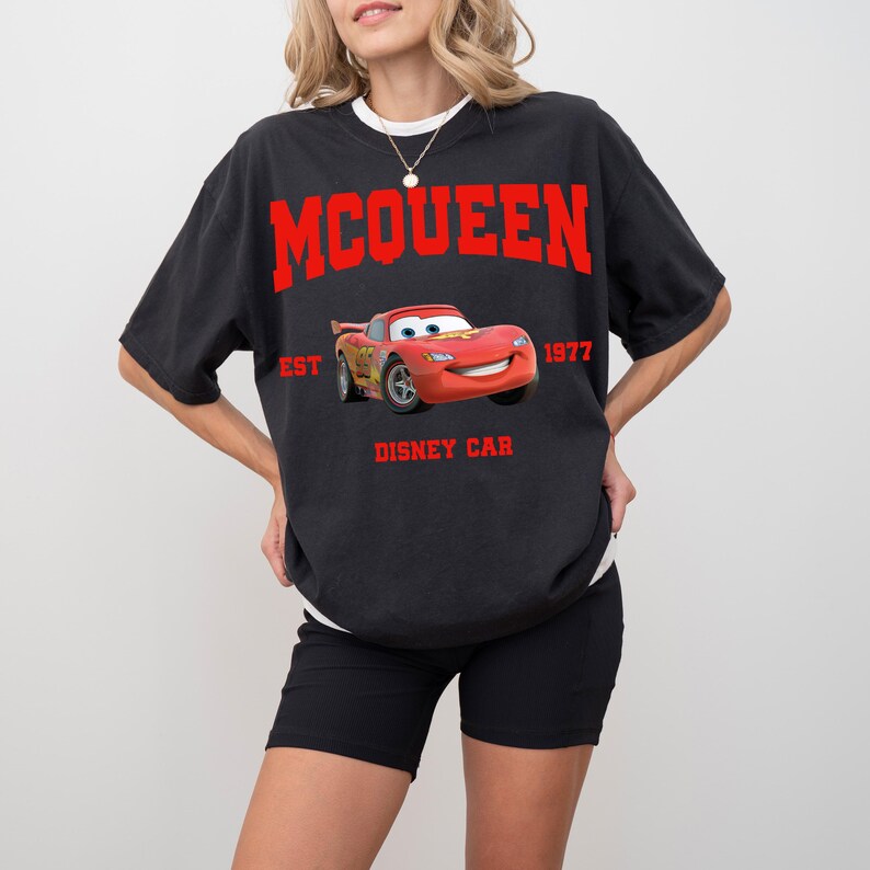 Peut inclure: T-shirt noir avec un motif de Flash McQueen du film Disney Cars. Le motif comprend le nom "McQueen" en lettres rouges, le texte "EST 1977" et "DISNEY CAR". Le t-shirt a un col rond et des manches courtes.