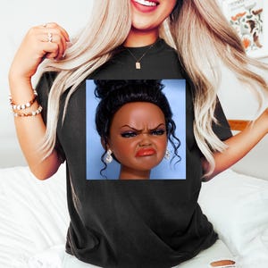 May include: Black t-shirt featuring a square image of a doll's head with a displeased expression. The doll has dark skin, dark hair, and is wearing earrings. The t-shirt is worn by a person with blonde hair and jewelry.