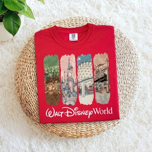 May include: Red crew neck t-shirt featuring four vertical rectangular panels with illustrations of Walt Disney World landmarks. The words "Walt Disney World" are printed in white script below the panels.