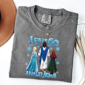 May include: Gray Comfort Colors t-shirt with the text "Let It Go and Let Jesus" and a graphic of Jesus, Elsa, and Anna. The shirt has a crew neck and short sleeves. A silver necklace with a pendant is on the shirt.