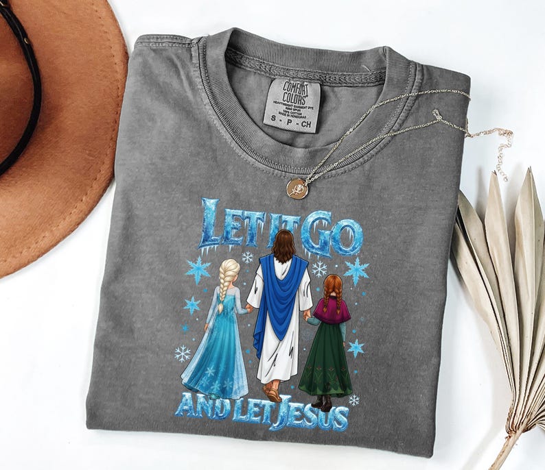 May include: Gray Comfort Colors t-shirt with the text "Let It Go and Let Jesus" and a graphic of Jesus, Elsa, and Anna. The shirt has a crew neck and short sleeves. A silver necklace with a pendant is on the shirt.