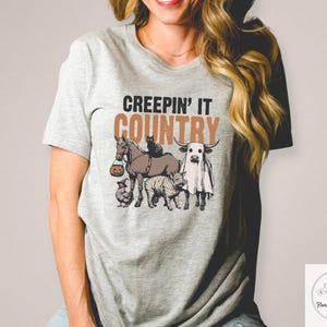 May include: A light gray t-shirt with the phrase "CREEPIN' IT COUNTRY" in brown and black text. The graphic features a horse, cat, cow, zebra, and chicken, all with Halloween-themed costumes.