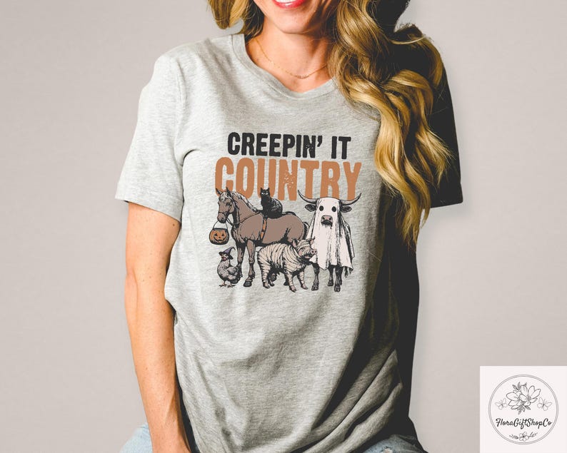 May include: A light gray t-shirt with the phrase "CREEPIN' IT COUNTRY" in brown and black text. The graphic features a horse, cat, cow, zebra, and chicken, all with Halloween-themed costumes.